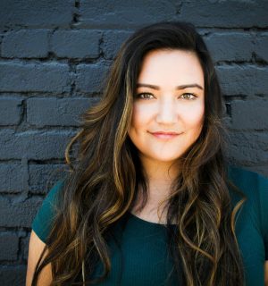 A captivating portrait of a woman smiling confidently in front of a brick wall, showcasing her long brunette hair and vibrant eyes.