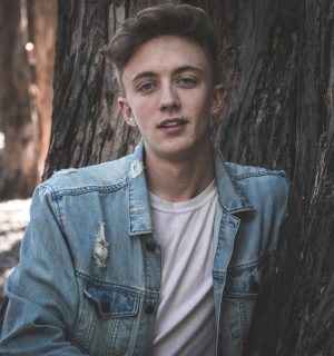Casual portrait of a young man in a denim jacket leaning against a tree.